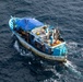 An aerial photograph of a small fishing vessel named 'MANTA' navigating open blue ocean waters. The boat displays a blue and white color scheme with visible wear and weathering on its hull. A tow line or fishing line extends from the stern into the water.