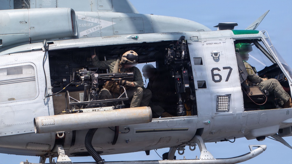 A close-up photograph of a military helicopter, numbered '67', with its side door open revealing armed crew members inside. At least two personnel are visible, one operating what appears to be a mounted multi-barrel rotary machine gun (likely an M134 minigun). The helicopter appears to be in flight or hovering, with warning markings visible including 'JET DANGER INTAKE' on the fuselage.