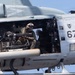 A close-up photograph of a military helicopter, numbered '67', with its side door open revealing armed crew members inside. At least two personnel are visible, one operating what appears to be a mounted multi-barrel rotary machine gun (likely an M134 minigun). The helicopter appears to be in flight or hovering, with warning markings visible including 'JET DANGER INTAKE' on the fuselage.