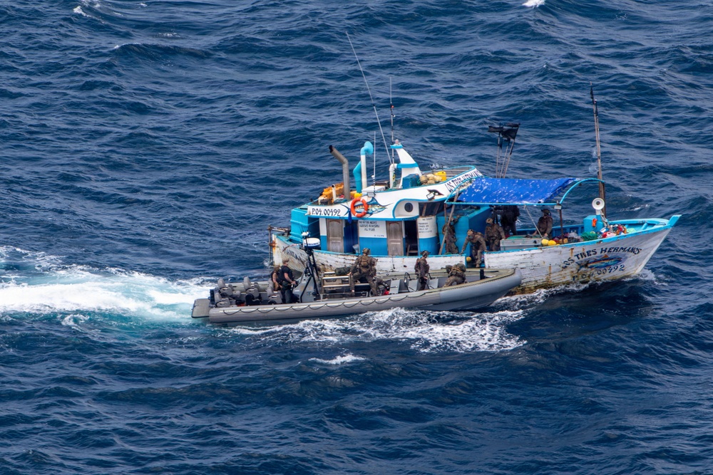 An aerial photograph shows a blue and white fishing vessel being boarded by what appear to be armed law enforcement or military personnel. A rigid inflatable boat (RIB) is alongside the larger vessel, with personnel in tactical gear visible on both craft. The vessel bears registration markings and the name 'Los Tres Hermanos' on its hull.