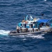 An aerial photograph shows a blue and white fishing vessel being boarded by what appear to be armed law enforcement or military personnel. A rigid inflatable boat (RIB) is alongside the larger vessel, with personnel in tactical gear visible on both craft. The vessel bears registration markings and the name 'Los Tres Hermanos' on its hull.