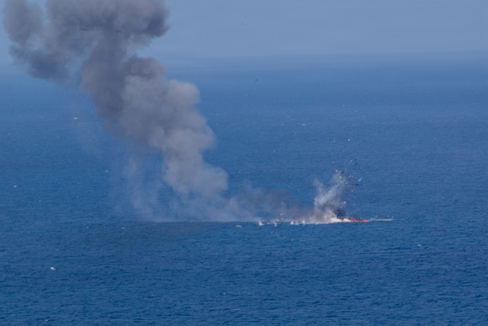 The image shows an explosion and fire occurring on the surface of open ocean water, producing a large column of dark gray smoke rising into the sky. Debris and sparks are visible being ejected from the point of ignition. The event appears to be a maritime incident, possibly a vessel sinking, controlled demolition exercise, or strike on a waterborne target.