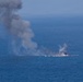 The image shows an explosion and fire occurring on the surface of open ocean water, producing a large column of dark gray smoke rising into the sky. Debris and sparks are visible being ejected from the point of ignition. The event appears to be a maritime incident, possibly a vessel sinking, controlled demolition exercise, or strike on a waterborne target.