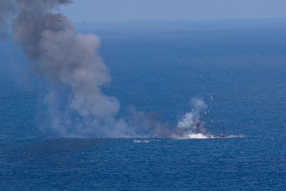 The image shows an explosion and fire occurring on the surface of open ocean water, producing a large column of dark gray smoke rising into the sky. Debris and white smoke are visible at the point of impact or detonation on the water's surface. The scene appears to depict a vessel or object being destroyed or sinking at sea.
