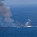 The image shows an explosion and fire occurring on the surface of open ocean water, producing a large column of dark gray smoke rising into the sky. Debris and white smoke are visible at the point of impact or detonation on the water's surface. The scene appears to depict a vessel or object being destroyed or sinking at sea.