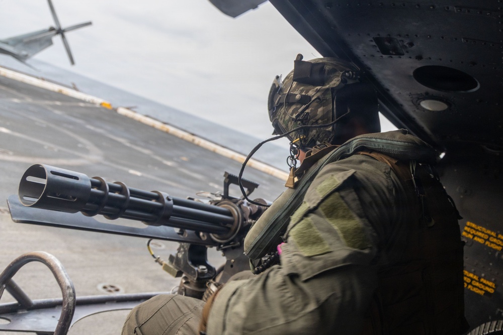 A military personnel is positioned at the open door of a helicopter, manning a multi-barrel rotary machine gun (consistent with an M134 Minigun). The operator is wearing camouflage combat gear, a tactical helmet with communications equipment, and is oriented toward what appears to be a ship deck or flight deck below. Yellow caution markings are partially visible on the helicopter's interior panel.