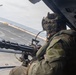 A military personnel is positioned at the open door of a helicopter, manning a multi-barrel rotary machine gun (consistent with an M134 Minigun). The operator is wearing camouflage combat gear, a tactical helmet with communications equipment, and is oriented toward what appears to be a ship deck or flight deck below. Yellow caution markings are partially visible on the helicopter's interior panel.
