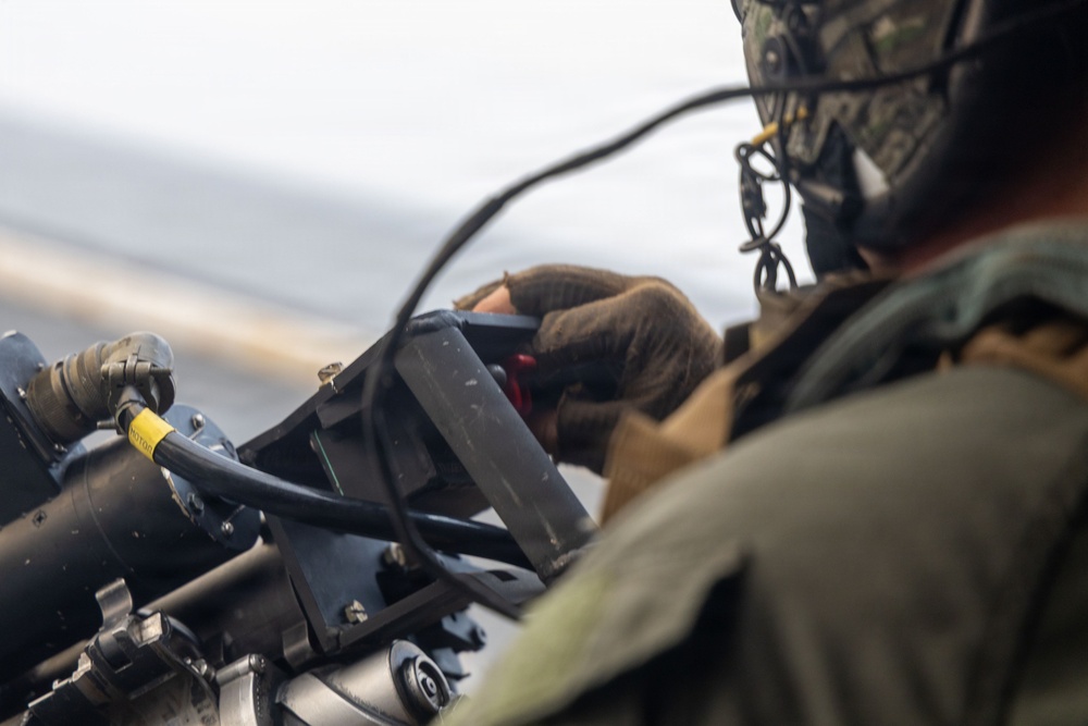 A military personnel wearing camouflage gear and a helmet is operating or adjusting a piece of equipment, likely a weapon system or sensor mount, in what appears to be an aerial or maritime environment. The individual is wearing tactical gloves and appears to be working with a mounted device that has cables and connectors attached. The background is blurred, suggesting motion or a wide aperture lens setting.