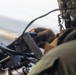A military personnel wearing camouflage gear and a helmet is operating or adjusting a piece of equipment, likely a weapon system or sensor mount, in what appears to be an aerial or maritime environment. The individual is wearing tactical gloves and appears to be working with a mounted device that has cables and connectors attached. The background is blurred, suggesting motion or a wide aperture lens setting.