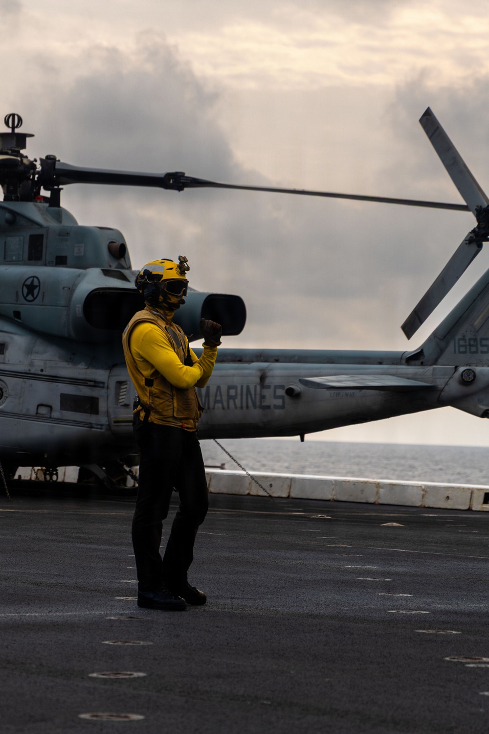 A flight deck crew member wearing a yellow jersey and protective gear stands on a naval vessel's flight deck, appearing to signal with a raised fist. Behind them is a U.S. Marines helicopter, identifiable by the 'MARINES' marking on its fuselage, chained to the deck. The scene takes place at sea under overcast skies.