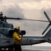 A flight deck crew member wearing a yellow jersey and protective gear stands on a naval vessel's flight deck, appearing to signal with a raised fist. Behind them is a U.S. Marines helicopter, identifiable by the 'MARINES' marking on its fuselage, chained to the deck. The scene takes place at sea under overcast skies.