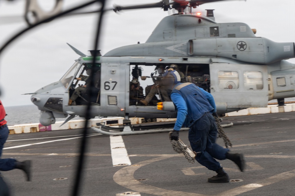 A military helicopter marked with the number '67' and a US military star insignia is on the flight deck of what appears to be a naval vessel. A crew member dressed in blue coveralls is running toward or away from the helicopter while carrying a chain, suggesting active deck operations. Additional personnel are visible inside the open cabin of the helicopter.