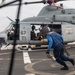 A military helicopter marked with the number '67' and a US military star insignia is on the flight deck of what appears to be a naval vessel. A crew member dressed in blue coveralls is running toward or away from the helicopter while carrying a chain, suggesting active deck operations. Additional personnel are visible inside the open cabin of the helicopter.