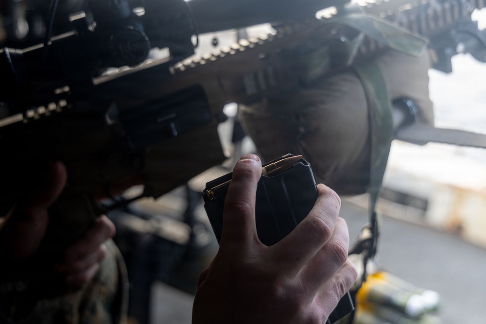A military or law enforcement personnel is holding a rifle magazine loaded with visible cartridges, positioned near a tactical firearm equipped with an optic sight and rail system. The image is taken at close range with a shallow depth of field, emphasizing the magazine and hand. The subject appears to be in the process of loading or inspecting the weapon.