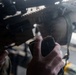 A military or law enforcement personnel is holding a rifle magazine loaded with visible cartridges, positioned near a tactical firearm equipped with an optic sight and rail system. The image is taken at close range with a shallow depth of field, emphasizing the magazine and hand. The subject appears to be in the process of loading or inspecting the weapon.