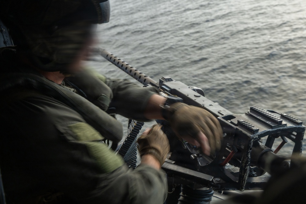 A military personnel member is operating a crew-served machine gun mounted on what appears to be a helicopter or aircraft door, positioned over open water. The operator is wearing flight gear including a helmet with communications equipment and olive drab flight suit. Motion blur on the hands suggests active weapon handling or loading.