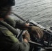 A military personnel member is operating a crew-served machine gun mounted on what appears to be a helicopter or aircraft door, positioned over open water. The operator is wearing flight gear including a helmet with communications equipment and olive drab flight suit. Motion blur on the hands suggests active weapon handling or loading.