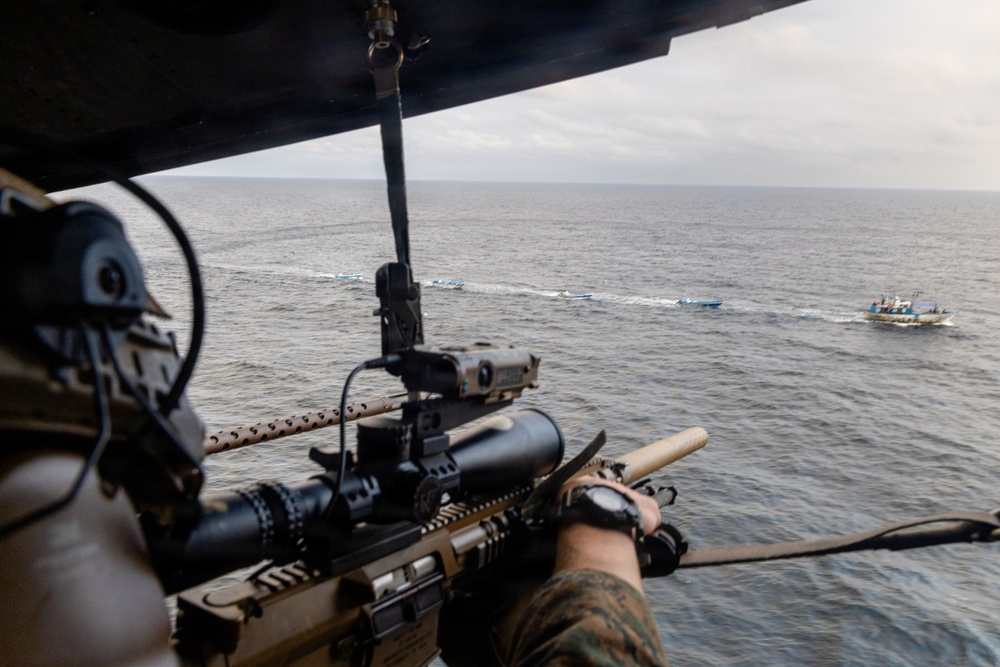 A military or law enforcement personnel is aiming a scoped rifle from an airborne helicopter toward small boats on the open ocean. The perspective is from inside the helicopter looking outward, with the shooter in the foreground and multiple small watercraft visible in the distance. The scene suggests an aerial interdiction or maritime surveillance operation.