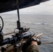 A military or law enforcement personnel is aiming a scoped rifle from an airborne helicopter toward small boats on the open ocean. The perspective is from inside the helicopter looking outward, with the shooter in the foreground and multiple small watercraft visible in the distance. The scene suggests an aerial interdiction or maritime surveillance operation.