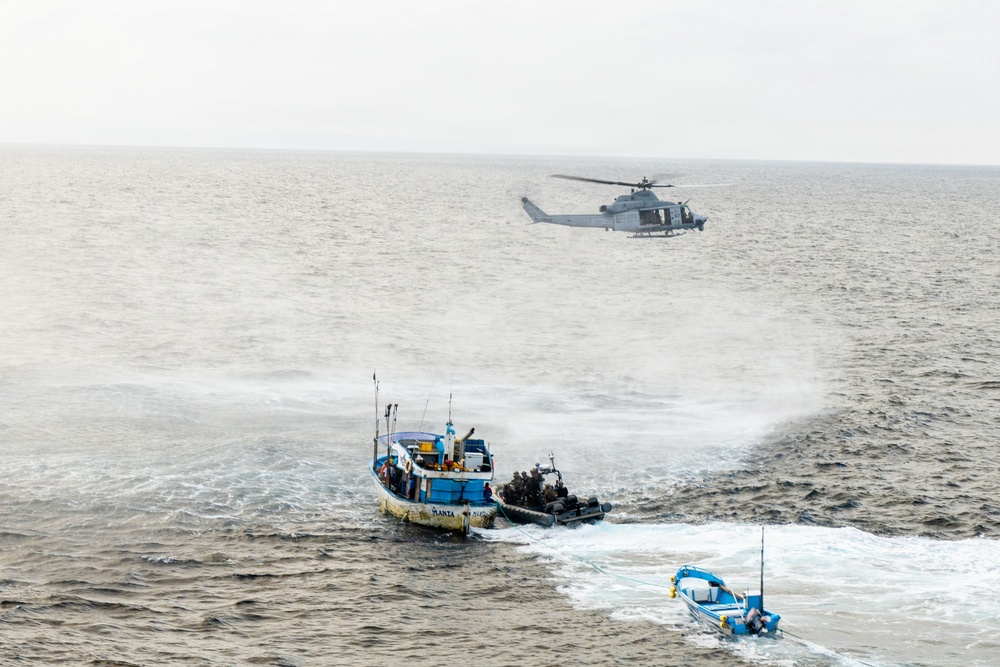 A maritime interdiction operation is underway at sea, with a military helicopter providing aerial support while a rigid inflatable boat (RIB) with armed personnel approaches a blue fishing vessel. A smaller blue boat is visible in the foreground being towed or secured. The scene depicts what appears to be a law enforcement or naval boarding operation in open ocean conditions.