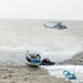 A maritime interdiction operation is underway at sea, with a military helicopter providing aerial support while a rigid inflatable boat (RIB) with armed personnel approaches a blue fishing vessel. A smaller blue boat is visible in the foreground being towed or secured. The scene depicts what appears to be a law enforcement or naval boarding operation in open ocean conditions.