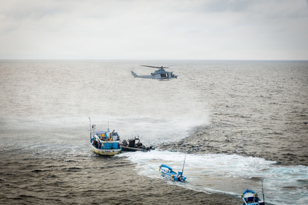 A military or law enforcement maritime interdiction operation is underway at sea. A gray military helicopter hovers over the water while a rigid inflatable boat (RIB) with armed personnel approaches a larger yellow and blue fishing vessel. Additional smaller blue boats are visible nearby, suggesting a coordinated multi-vessel operation.