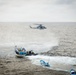 A military or law enforcement maritime interdiction operation is underway at sea. A gray military helicopter hovers over the water while a rigid inflatable boat (RIB) with armed personnel approaches a larger yellow and blue fishing vessel. Additional smaller blue boats are visible nearby, suggesting a coordinated multi-vessel operation.