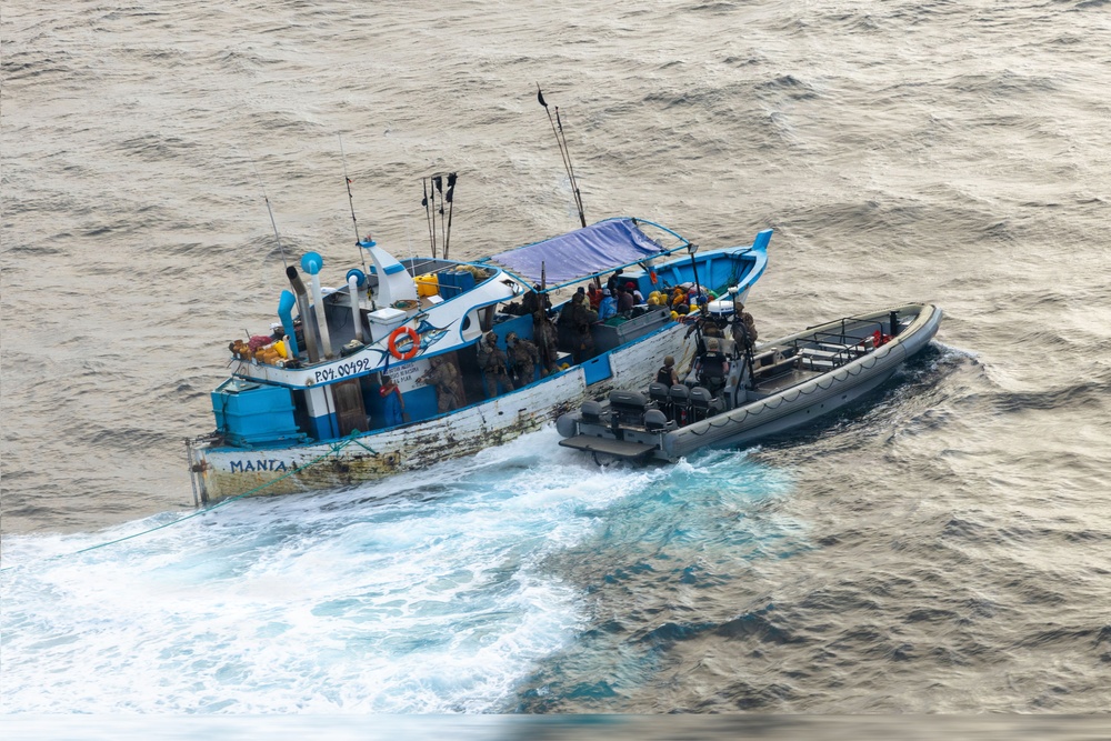 An aerial photograph shows a gray rigid inflatable boat (RIB) alongside a weathered blue-and-white fishing vessel named 'MANTA I' at sea. Multiple personnel in tactical/military gear are visible on both vessels, suggesting a maritime interdiction or boarding operation. The fishing vessel shows significant wear and carries a blue tarpaulin canopy over its deck.