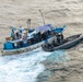 An aerial photograph shows a gray rigid inflatable boat (RIB) alongside a weathered blue-and-white fishing vessel named 'MANTA I' at sea. Multiple personnel in tactical/military gear are visible on both vessels, suggesting a maritime interdiction or boarding operation. The fishing vessel shows significant wear and carries a blue tarpaulin canopy over its deck.