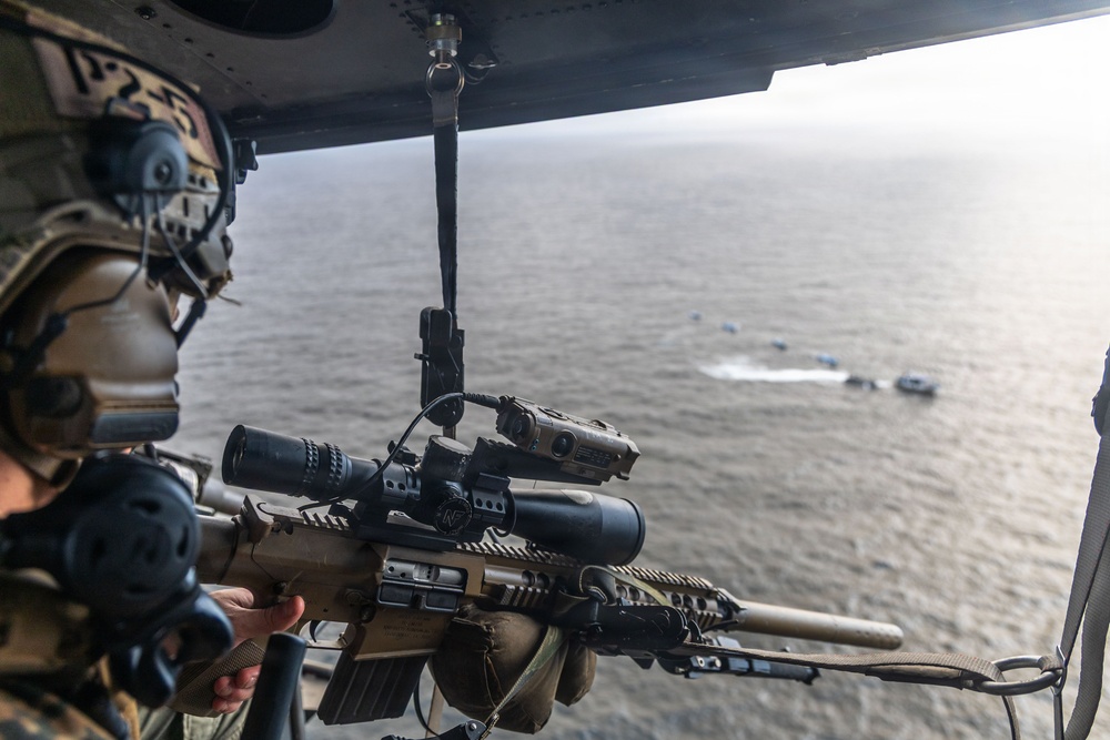 A military operator is positioned at the open door of a helicopter, holding a scoped rifle with multiple attachments while observing a maritime scene below. Several small watercraft are visible on the ocean surface in the background. The image appears to document an aerial maritime surveillance or overwatch operation.