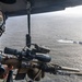 A military operator is positioned at the open door of a helicopter, holding a scoped rifle with multiple attachments while observing a maritime scene below. Several small watercraft are visible on the ocean surface in the background. The image appears to document an aerial maritime surveillance or overwatch operation.
