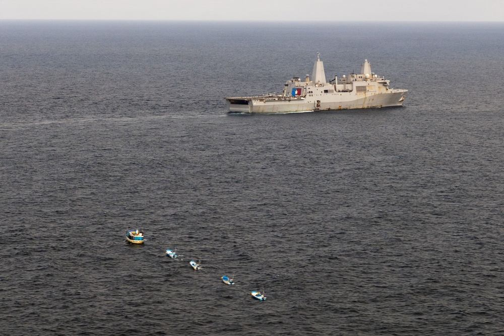An aerial photograph shows a large gray naval vessel, likely a US Navy amphibious transport dock ship, underway on open ocean. In the foreground, five small civilian-style boats are arranged in a loose diagonal line on the water. The scene suggests a maritime encounter or patrol situation between the warship and the smaller vessels.