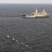 An aerial photograph shows a large gray naval vessel, likely a US Navy amphibious transport dock ship, underway on open ocean. In the foreground, five small civilian-style boats are arranged in a loose diagonal line on the water. The scene suggests a maritime encounter or patrol situation between the warship and the smaller vessels.