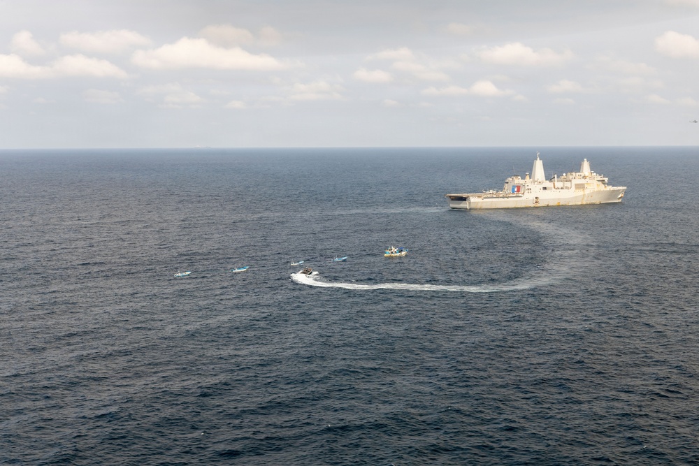An aerial photograph shows a large naval warship, appearing to be a US Navy amphibious transport dock vessel, operating at sea alongside several smaller rigid inflatable boats (RIBs) and small craft. The smaller vessels are arranged in a loose formation near the larger ship, with at least one boat leaving a visible wake from a turning maneuver. The scene appears to depict a maritime exercise, small boat operations, or a boarding/interdiction drill.