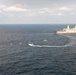 An aerial photograph shows a large naval warship, appearing to be a US Navy amphibious transport dock vessel, operating at sea alongside several smaller rigid inflatable boats (RIBs) and small craft. The smaller vessels are arranged in a loose formation near the larger ship, with at least one boat leaving a visible wake from a turning maneuver. The scene appears to depict a maritime exercise, small boat operations, or a boarding/interdiction drill.