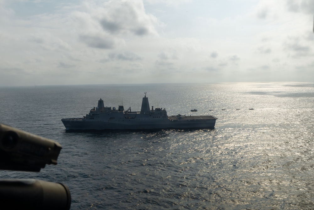 An aerial photograph taken from a military aircraft showing a large naval warship, consistent with a San Antonio-class amphibious transport dock (LPD), underway on open ocean. The image appears to be taken from a helicopter or aircraft, with part of the aircraft's fuselage visible in the lower left foreground. Several smaller vessels are visible in the distance to the right of the main ship.