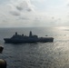 An aerial photograph taken from a military aircraft showing a large naval warship, consistent with a San Antonio-class amphibious transport dock (LPD), underway on open ocean. The image appears to be taken from a helicopter or aircraft, with part of the aircraft's fuselage visible in the lower left foreground. Several smaller vessels are visible in the distance to the right of the main ship.