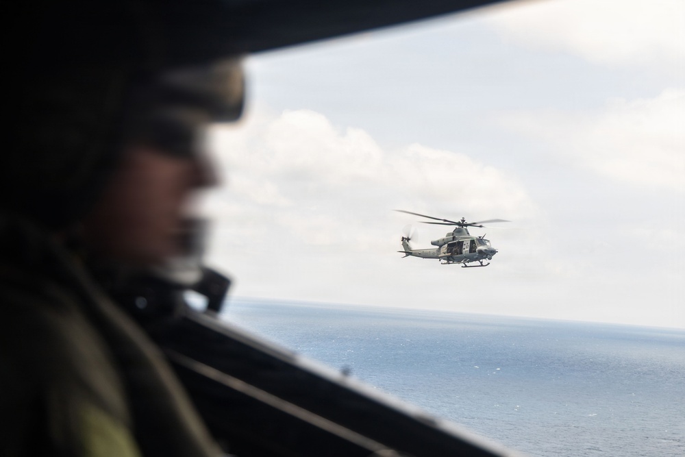 A military personnel member wearing a flight helmet is visible in the foreground, photographed from inside an aircraft looking out a window. In the background, a military helicopter — consistent with a UH-1Y Venom or similar twin-engine utility helicopter — flies over open ocean water. The image captures a formation or escort flight scenario over a calm sea under partly cloudy skies.