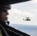 A military personnel member wearing a flight helmet is visible in the foreground, photographed from inside an aircraft looking out a window. In the background, a military helicopter — consistent with a UH-1Y Venom or similar twin-engine utility helicopter — flies over open ocean water. The image captures a formation or escort flight scenario over a calm sea under partly cloudy skies.
