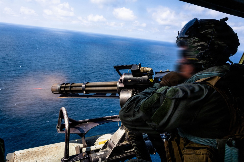 A military operator is firing a multi-barrel rotary machine gun (consistent with an M134 Minigun) from what appears to be the open door of a helicopter over open ocean. Muzzle flash is visible at the barrel end, indicating active firing. The operator is wearing full tactical gear including a flight helmet with communications equipment.