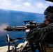 A military operator is firing a multi-barrel rotary machine gun (consistent with an M134 Minigun) from what appears to be the open door of a helicopter over open ocean. Muzzle flash is visible at the barrel end, indicating active firing. The operator is wearing full tactical gear including a flight helmet with communications equipment.