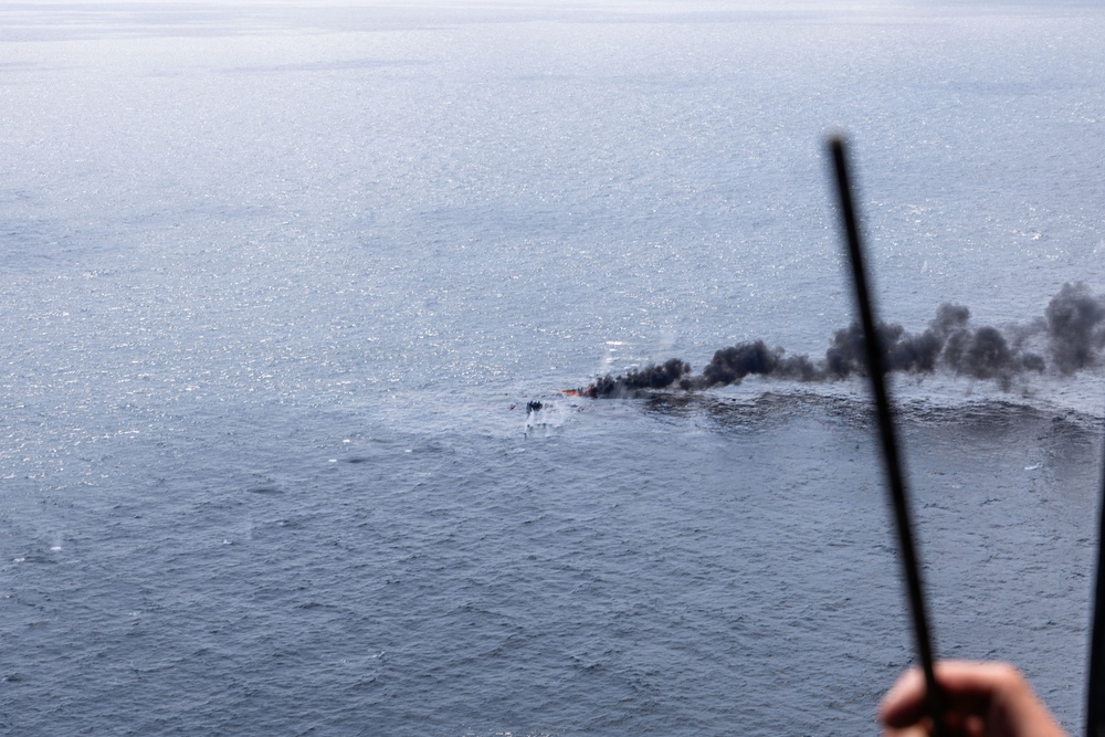 An aerial photograph taken from an elevated platform or aircraft showing a burning object on the open ocean surface. Dark black smoke billows from what appears to be a small burning vessel or debris in the water. The photo is taken from a significant height, with a rod or antenna visible in the foreground.