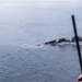 An aerial photograph taken from an elevated platform or aircraft showing a burning object on the open ocean surface. Dark black smoke billows from what appears to be a small burning vessel or debris in the water. The photo is taken from a significant height, with a rod or antenna visible in the foreground.