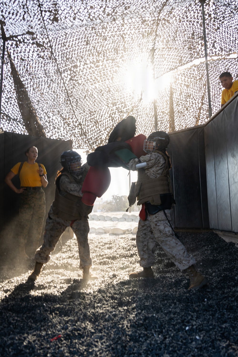 Two individuals in military camouflage uniforms and protective gear (helmets and padding) are engaged in a close-quarters combat training exercise under a camouflage netting structure. One participant is executing a knee strike while the other defends with padded arm guards. Observers in yellow shirts and additional personnel watch from the periphery, suggesting an instructional or evaluation setting.