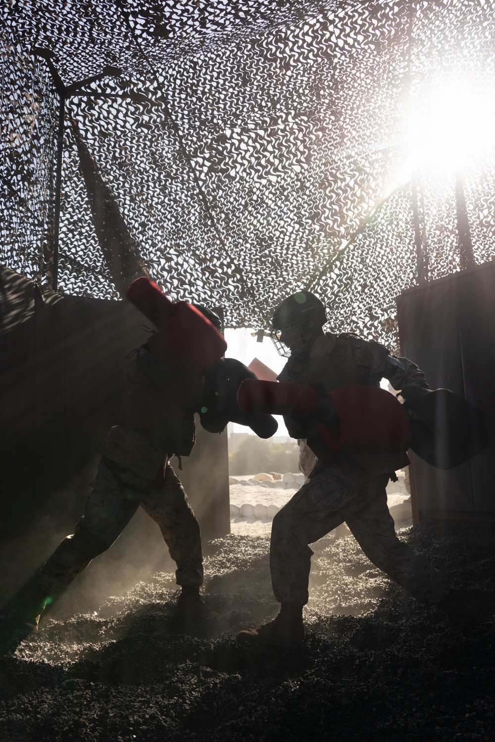 Two individuals in military camouflage uniforms are engaged in a combatives or boxing training exercise under a camouflage netting structure. One participant is wearing a protective face cage helmet and both are using large red padded boxing/sparring gloves. The scene is dramatically backlit by sunlight filtering through the netting, creating silhouettes and dust particles in the air.