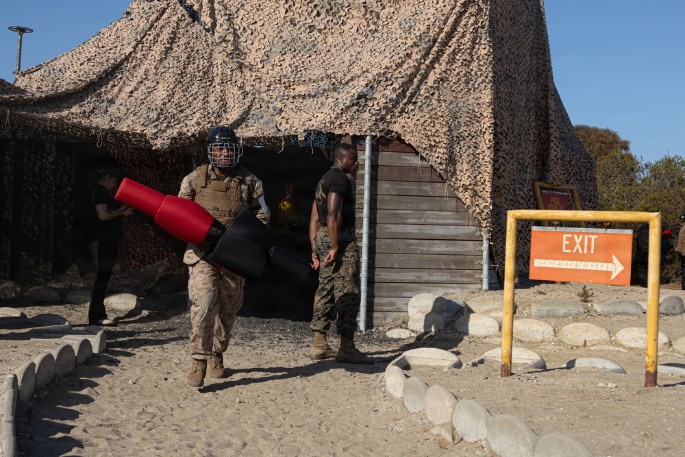 The image shows military personnel, likely U.S. Marines based on their camouflage uniforms, at what appears to be a training facility or obstacle course. One service member is wearing a protective helmet and body armor while carrying padded training equipment (red and black foam/padding). The setting features a camouflage netting structure, wooden walls, and an orange EXIT sign with an arrow.