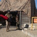 The image shows military personnel, likely U.S. Marines based on their camouflage uniforms, at what appears to be a training facility or obstacle course. One service member is wearing a protective helmet and body armor while carrying padded training equipment (red and black foam/padding). The setting features a camouflage netting structure, wooden walls, and an orange EXIT sign with an arrow.