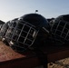 Multiple black Bauer hockey helmets with wire cage face guards are lined up on what appears to be a metal railing or bench. The helmets are photographed in an outdoor setting with natural backlighting creating a dramatic silhouette effect. The image appears to be taken at a hockey facility or outdoor rink area.