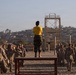 A military drill instructor wearing a yellow 'CDI' shirt stands elevated on a table, leading a large group of recruits in physical training exercises on a sandy outdoor area. The recruits are dressed in camouflage uniforms and appear to be performing synchronized movements. The setting appears to be a military training base, likely MCRD (Marine Corps Recruit Depot) San Diego, based on the visible infrastructure and coastal hillside backdrop.