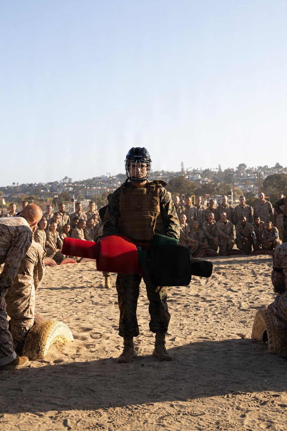 A military training exercise is taking place on a sandy outdoor area, likely a Marine Corps base given the MARPAT camouflage uniforms visible throughout. A central figure wearing a Bauer hockey-style helmet with face cage, plate carrier vest, and camouflage uniform holds padded training equipment (red and black striking pads). A large group of uniformed personnel observes from the background in a semicircle formation.
